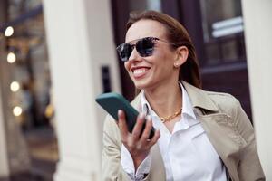 sorridente europeo donna nel occhiali da sole è in piedi con Telefono su città strada sfondo foto