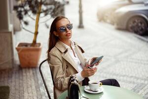 bellissimo donna indossare occhiali da sole seduta a bar terrazza Tenere Telefono mentre potabile caffè foto
