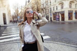 attraente alla moda donna nel cappotto attraversamento il città strada e guardare a lato foto