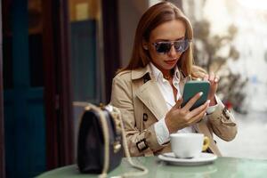 attraente elegante donna nel bicchieri seduta a bar terrazza e uso Telefono mentre potabile caffè foto