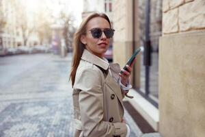 sorridente europeo donna nel occhiali da sole è in piedi con Telefono su città strada sfondo foto
