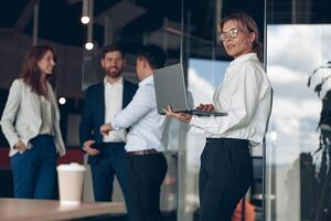 maturo fiducioso donna d'affari a ufficio con gruppo di colleghi su sfondo, Lavorando su il computer portatile foto