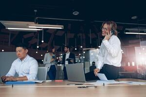 sorridente attività commerciale donna seduta su tavolo e parlando su Telefono dopo incontro nel moderno ufficio foto