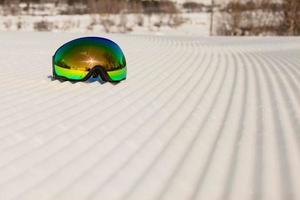 occhiali da sci posati su una nuova neve battuta e una pista da sci vuota foto