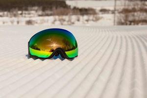 occhiali da sci posati su una nuova neve battuta e una pista da sci vuota foto