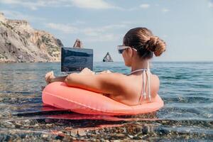 donna libero professionista lavori su il computer portatile nuoto nel mare su rosa gonfiabile squillo. contento turista nel occhiali da sole galleggiante su gonfiabile ciambella e Lavorando su il computer portatile computer nel calma oceano. a distanza Lavorando dovunque foto