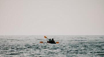 contento coppia kayak nel un gonfiabile kayak su il mare a tramonto. coppia canoa nel il mare vicino il isola con montagne. persone kayak nel vita giacche vela. indietro Visualizza foto