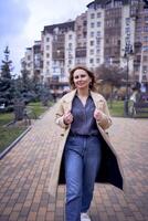 giovane adulto donna nel jeans e sabbia cappotto passeggiate attraverso il città su un' primavera giorno, moderno architettura dietro a foto