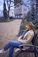 giovane adulto donna nel jeans e sabbia cappotto seduta su un' panchina su un' primavera giorno, moderno architettura dietro a foto
