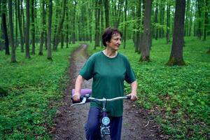 anziano donna a piedi nel primavera foresta con bicicletta e yoga stuoia foto