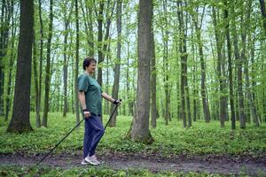 anziano donna è impegnato nel nordico a piedi con bastoni nel il primavera foresta foto