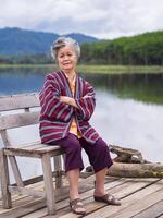 ritratto di un anziano asiatico donna sorridente e braccia attraversato e guardare a il telecamera mentre seduta lato il lago. concetto di anziano persone e rilassamento foto