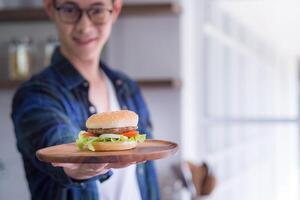 giovane uomo indossare bicchieri sorridente, guardare a il telecamera, e Tenere un' fatti in casa Hamburger su un' di legno piatto con sfocatura sfondo. spazio per testo. selettivo messa a fuoco. concetto di cucinando foto