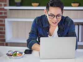 un' giovane uomo è eccitato di un' nuovo progetto. uomo d'affari seduta nel il ufficio, lettura un' rapporto a partire dal un' programma su il il computer portatile e sorridente molto contento. Lavorando con prima colazione. picco ore foto