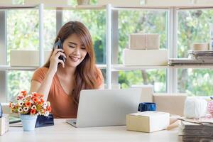 asiatico donna d'affari utilizzando un' smartphone e guardare a un' il computer portatile mentre seduta nel il ufficio. giovane donna di negoziazione di attività commerciale. concetto di attività commerciale e e-commerce foto