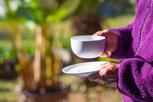 avvicinamento di mani anziano donna Tenere un' bianca caffè tazza mentre in piedi nel un' giardino. concetto di anziano persone e rilassamento foto