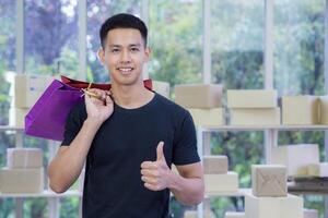 asiatico uomo d'affari in piedi Tenere colorato shopping borse e dando pollice su mentre in piedi nel il ufficio. spazio per testo. concetto di attività commerciale e e-commerce foto