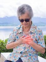 un' anziano donna stringendo sua il petto nel dolore a il segni di angina o miocardico infarto o cuore attacco mentre seduta a il lato di il lago. concetto di anziano persone e cuore attacco foto