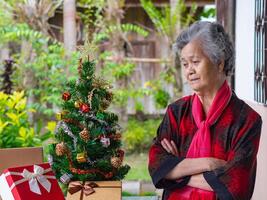 ritratto di anziano asiatico donna braccia attraversato e guardare a il Natale albero mentre in piedi a casa. concetto di anziano persone e festival foto