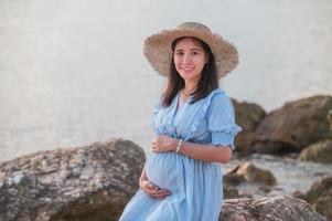 donna incinta seduta sulle rocce vicino alla spiaggia di sabbia e al mare. foto