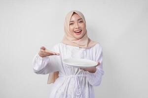 sorridente giovane asiatico musulmano donna nel un' velo hijab puntamento per un vuoto piatto con copia spazio al di sopra di esso, presentazione cibo menù per iftar. Ramadan concetto foto