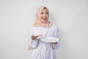 sorridente giovane asiatico musulmano donna nel un' velo hijab puntamento per un vuoto piatto con copia spazio al di sopra di esso, presentazione cibo menù per iftar. Ramadan concetto foto