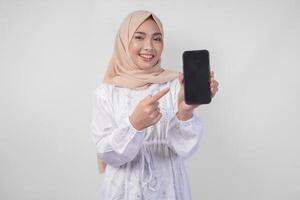 ritratto di giovane asiatico musulmano donna indossare bianca vestito e hijab sorridente felicemente, puntamento per il vuoto schermo copia spazio su sua Telefono e mostrando schermo per il telecamera foto