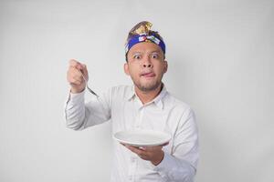 Affamato giovane balinese uomo nel bianca camicia e tradizionale copricapo Tenere un vuoto piatto con copia spazio e cucchiaio mentre fabbricazione un' divertente espressione foto