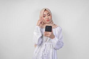 riflessivo giovane asiatico musulmano donna indossare bianca vestito e hijab, utilizzando smartphone mentre Tenere sua mento e pensiero con grave espressione al di sopra di isolato bianca sfondo foto