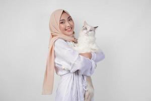 ritratto di un' bellissimo giovane musulmano donna indossare bianca vestito e hijab, sorridente e Tenere un' bianca bambola di pezza gatto al di sopra di isolato bianca sfondo foto