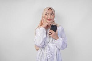 riflessivo giovane asiatico musulmano donna indossare bianca vestito e hijab, utilizzando smartphone mentre Tenere sua mento e pensiero con grave espressione al di sopra di isolato bianca sfondo foto