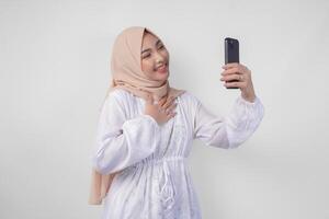 ritratto di asiatico musulmano donna indossare bianca vestito e hijab Tenere sua smartphone per fare un' chiamata per famiglia o amico, isolato di bianca sfondo. Ramadhan e eid mubarak concetto foto