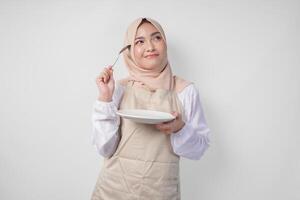 confuso giovane asiatico musulmano donna nel hijab e crema grembiule Tenere cucchiaio e vuoto piatto con copia spazio al di sopra di esso, pensiero difficile che cosa cibo menù per mangiare per iftar. Ramadan concetto foto