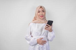 ritratto di giovane asiatico musulmano donna nel bianca vestito e hijab sensazione Affamato mentre guardare a sua Telefono per ordine delizioso cibo dopo digiuno, isolato su bianca sfondo foto