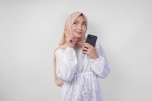 riflessivo giovane asiatico musulmano donna indossare bianca vestito e hijab, utilizzando smartphone mentre Tenere sua mento e pensiero con grave espressione al di sopra di isolato bianca sfondo foto