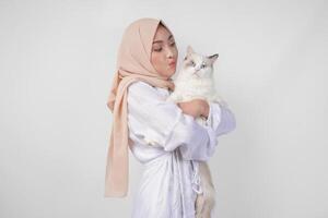 ritratto di un' bellissimo giovane musulmano donna indossare bianca vestito e hijab, sorridente e Tenere un' bianca bambola di pezza gatto al di sopra di isolato bianca sfondo foto