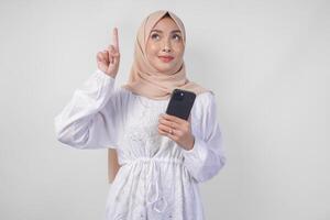 sorridente asiatico donna indossare hijab e bianca vestito utilizzando smartphone mentre puntamento su per il copia spazio sopra, in piedi al di sopra di isolato bianca sfondo. Ramadan e eid mubarak annuncio pubblicitario concetto foto