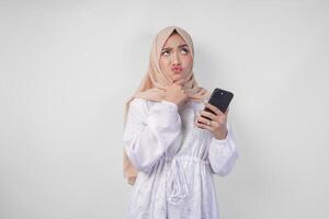 riflessivo giovane asiatico musulmano donna indossare bianca vestito e hijab, utilizzando smartphone mentre Tenere sua mento e pensiero con grave espressione al di sopra di isolato bianca sfondo foto
