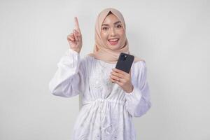 sorridente asiatico donna indossare hijab e bianca vestito utilizzando smartphone mentre puntamento su per il copia spazio sopra, in piedi al di sopra di isolato bianca sfondo. Ramadan e eid mubarak annuncio pubblicitario concetto foto