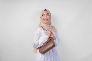 elegante giovane asiatico musulmano donna indossare bianca vestito e hijab con Marrone fionda Borsa sorridente felicemente al di sopra di isolato bianca sfondo. Ramadhan moda concetto foto