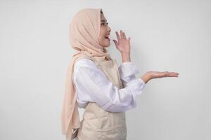 eccitato giovane asiatico musulmano donna nel hijab e crema grembiule puntamento per il vuoto copia spazio su il sinistra e giusto lato. Ramadan annuncio pubblicitario concetto foto