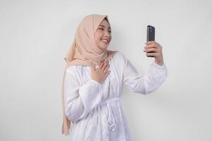 ritratto di asiatico musulmano donna indossare bianca vestito e hijab Tenere sua smartphone per fare un' chiamata per famiglia o amico, isolato di bianca sfondo. Ramadhan e eid mubarak concetto foto