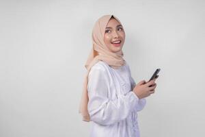riflessivo giovane asiatico musulmano donna indossare bianca vestito e hijab Tenere Telefono mentre pensiero e scoperta idea con contento espressione, isolato su bianca sfondo. Ramadan e eid mubarak concetto foto