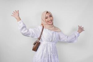 elegante giovane asiatico musulmano donna indossare bianca vestito e hijab con Marrone fionda Borsa sorridente felicemente al di sopra di isolato bianca sfondo. Ramadhan moda concetto foto