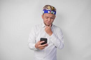 grave giovane balinese uomo nel bianca camicia e tradizionale copricapo Tenere smartphone mentre pensiero difficile foto