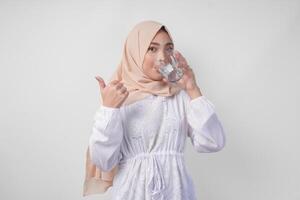 assetato giovane asiatico musulmano donna nel bianca vestito e crema velo hijab potabile fresco minerale acqua dopo digiuno mentre fare pollici su gesto. Ramadan concetto foto