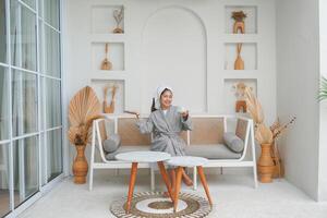sorridente giovane asiatico ragazza indossare grigio accappatoio potabile tazza di tè e sinistra mano Tenere copia spazio immaginario su il spazio foto