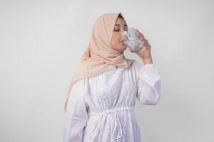 assetato giovane asiatico musulmano donna nel bianca vestito e crema velo hijab potabile fresco minerale acqua dopo digiuno. Ramadan concetto foto