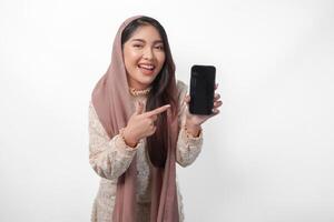 eccitato giovane asiatico musulmano donna nel foulard velo hijab puntamento per il copia spazio a smartphone vuoto schermo, isolato su bianca sfondo studio. Ramadan e eid mubarak concetto. foto