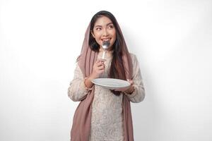 Affamato asiatico musulmano donna Tenere piatto pensiero che cosa per mangiare con copia spazio al di sopra di il piatto mentre pungente cucchiaio in attesa per iftar, isolato bianca sfondo foto